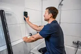A person uses a wall detector on white bathroom tiles.