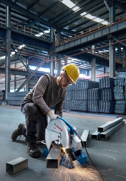 A person wearing safety equipment cuts metal tubing with a chop saw in a warehouse.