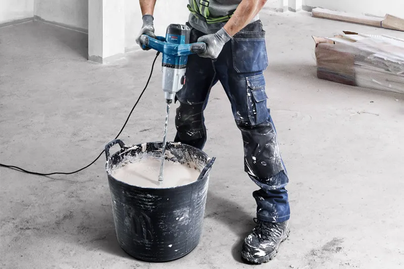 A person wearing safety equipment uses a stirring mechanism to mix material in a large bucket.