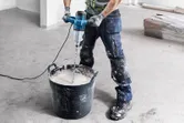 A person wearing safety equipment uses a stirring mechanism to mix material in a large bucket.