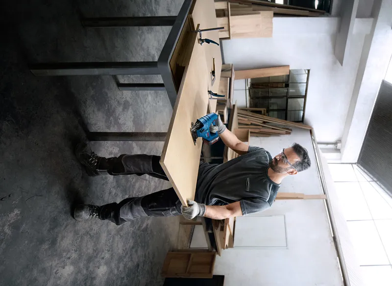 A person wearing safety equipment cuts wood with a power tool in a workshop.
