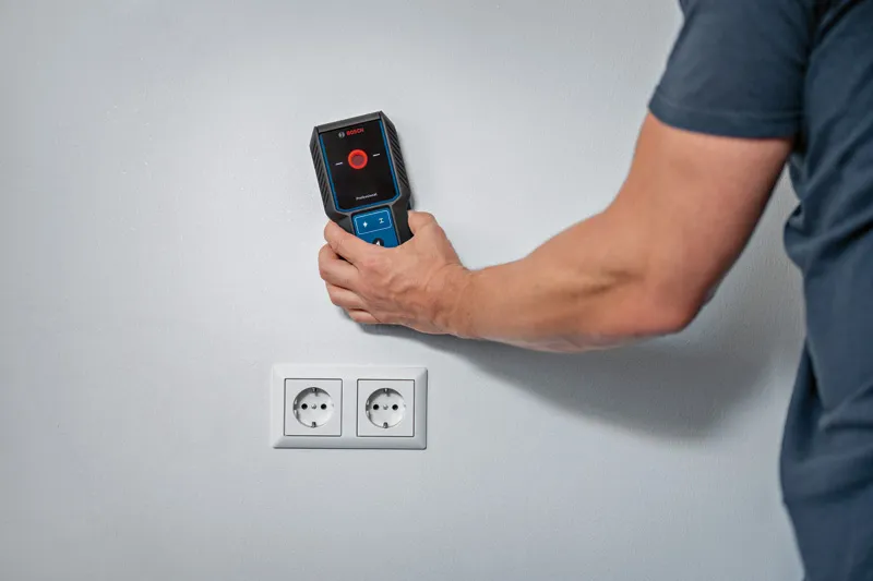 Person uses a wall detector above electrical outlets on a white wall.