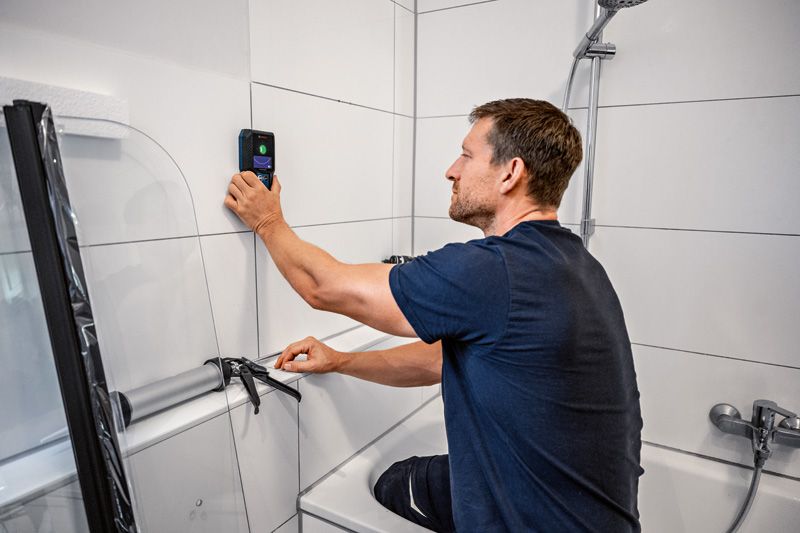 A person uses a detector on a tiled bathroom wall while preparing to apply sealant.