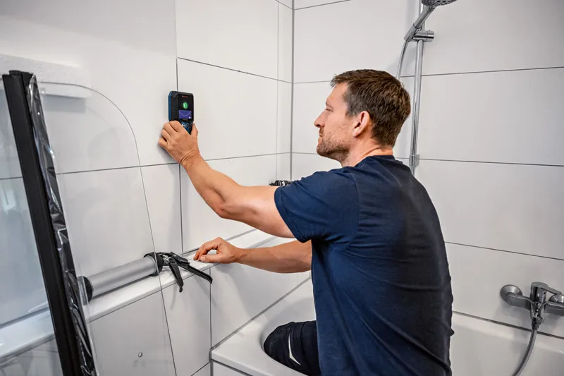 A person uses a detector on a tiled bathroom wall while preparing to apply sealant.