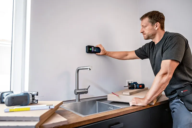 A person scans a kitchen wall with a detector above a sink.