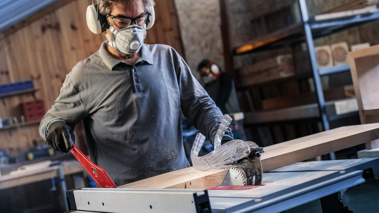 A person wearing safety equipment cuts a wooden board with a table saw.