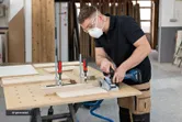 A person wearing safety equipment uses a biscuit joiner on clamped wood pieces.