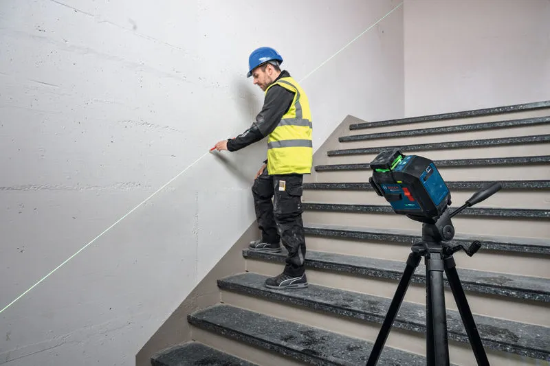 A person wearing safety equipment marks a wall on stairs using a laser leveling tool.