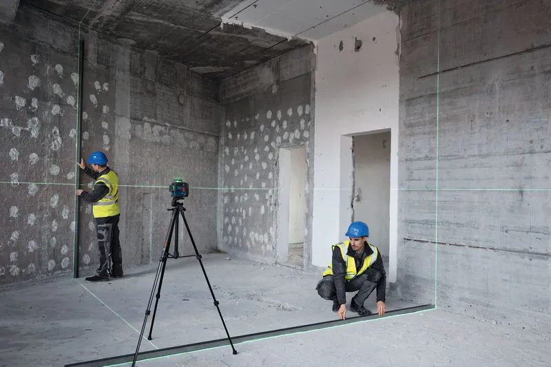 Two persons wearing safety equipment align a room with a laser leveling tool on a tripod.
