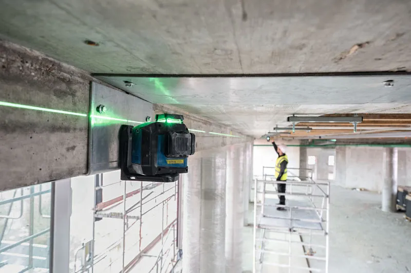 Laser leveling tool projects a green line across a concrete beam as a person wearing safety equipment works nearby.