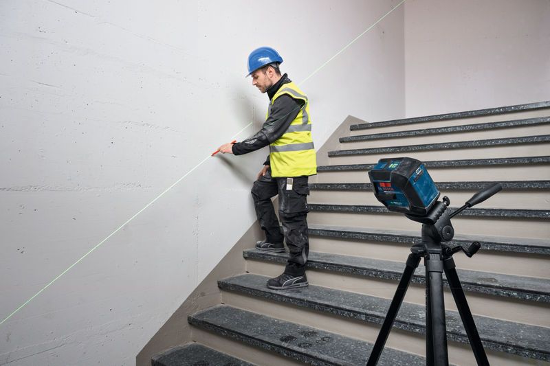 A person wearing safety equipment marks a stairwell wall using a laser leveling tool.