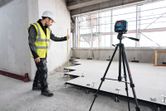 Person wearing safety equipment measures floor height using a laser leveling tool on a tripod.