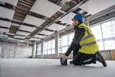 A person wearing safety equipment positions a laser leveling tool on the floor of a construction site.
