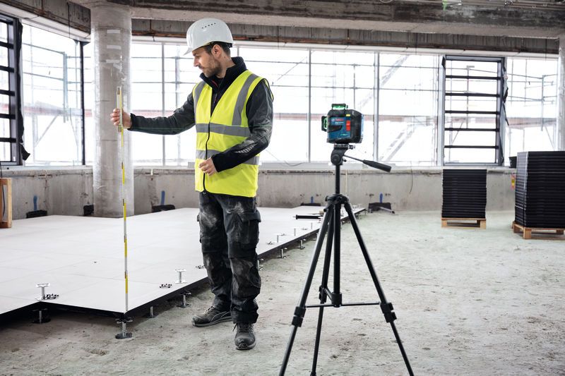 A person wearing safety equipment uses a laser leveling tool on a tripod while measuring floor height.