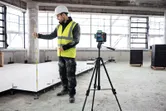 A person wearing safety equipment uses a laser leveling tool on a tripod while measuring floor height.