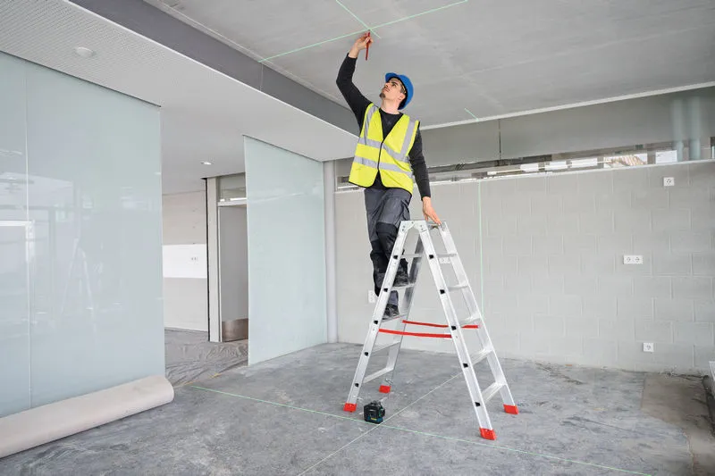 A person wearing safety equipment marks a ceiling using a laser leveling tool.