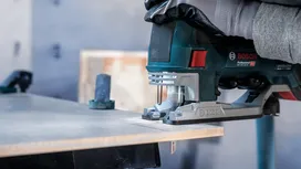 Person wearing safety equipment cuts wood with a power jigsaw.