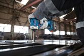 A person wearing safety equipment uses a cordless impact driver on a metal surface.