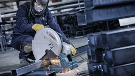 A person wearing safety equipment cuts metal beams with a power saw, producing sparks.
