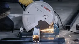 Person wearing safety equipment cuts a metal beam with a chop saw, producing sparks.