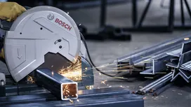 Person wearing safety equipment cuts steel tubing with a metal cut-off saw, sparks flying.