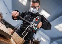 Person wearing safety equipment drills into wood indoors.