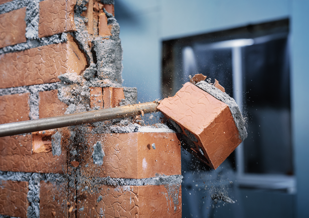 A demolition tool breaks a brick wall, scattering debris.