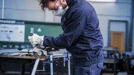 A person wearing safety equipment uses an angle grinder to cut metal, producing sparks.