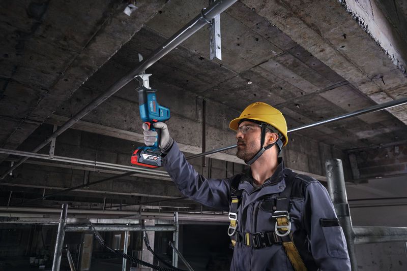 A worker wearing safety equipment uses a reciprocating saw to cut a ceiling pipe.