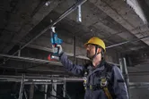A worker wearing safety equipment uses a reciprocating saw to cut a ceiling pipe.