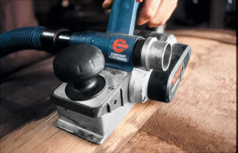 A person uses an electric planer with dust extraction to smooth a wooden surface.