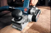 A person uses an electric planer with dust extraction to smooth a wooden surface.