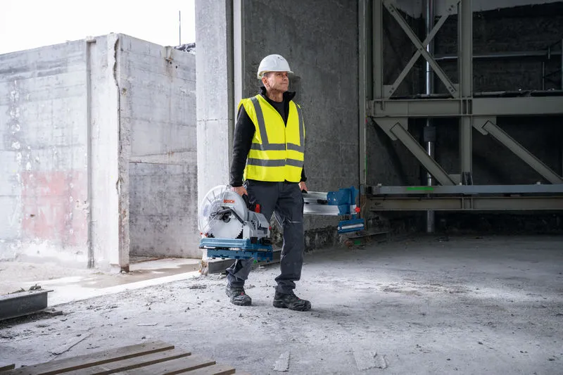A person wearing safety equipment carries a metal cut-off saw in an unfinished building.