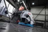 A person wearing safety equipment prepares a metal cut-off saw to cut a metal pipe.