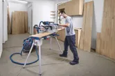 A person wearing safety equipment cuts a wooden board using a mitre saw in a workshop.