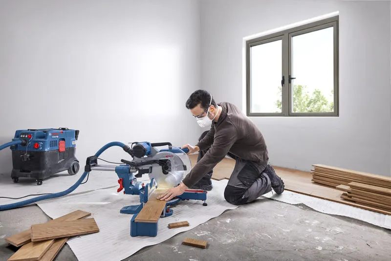 A person wearing safety equipment cuts wood planks with a mitre saw in a bright room.