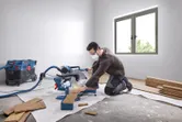 A person wearing safety equipment cuts wood planks with a mitre saw in a bright room.