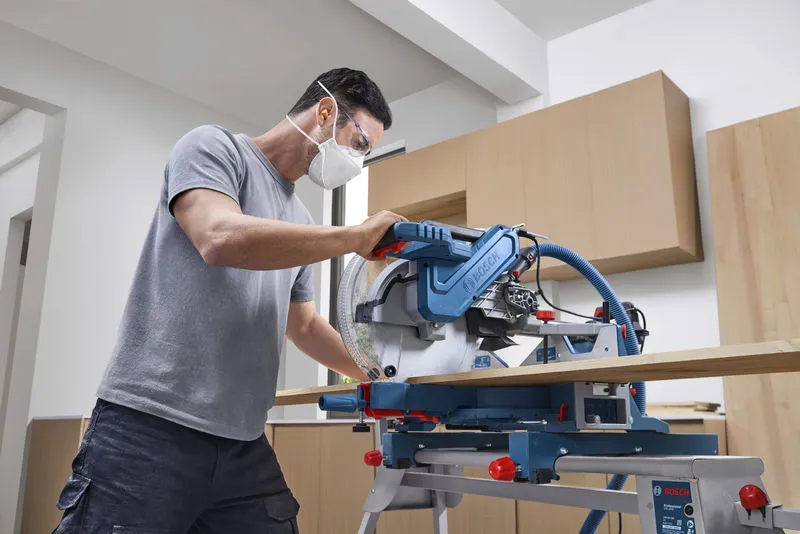 A person wearing safety equipment cuts wood with a mitre saw in a workshop.
