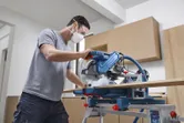 A person wearing safety equipment cuts wood with a mitre saw in a workshop.
