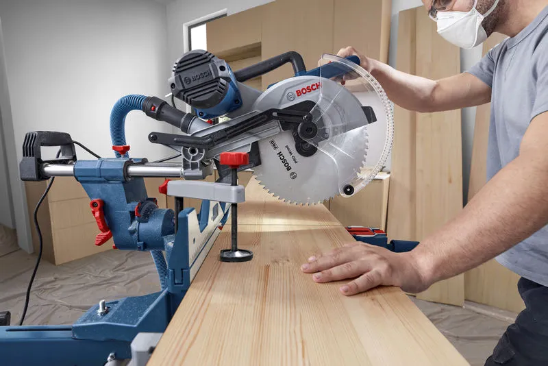 A person operates a mitre saw to cut a wooden board in a workshop.