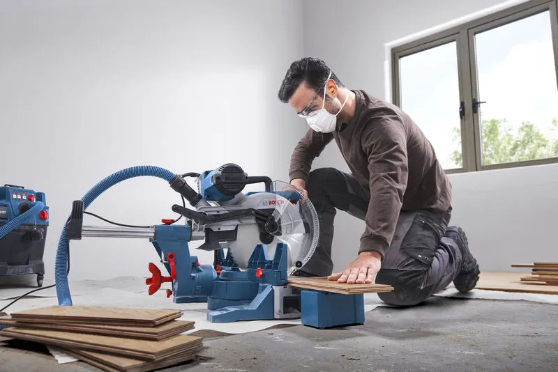 A person wearing safety equipment cuts wooden flooring with a mitre saw in a bright room.