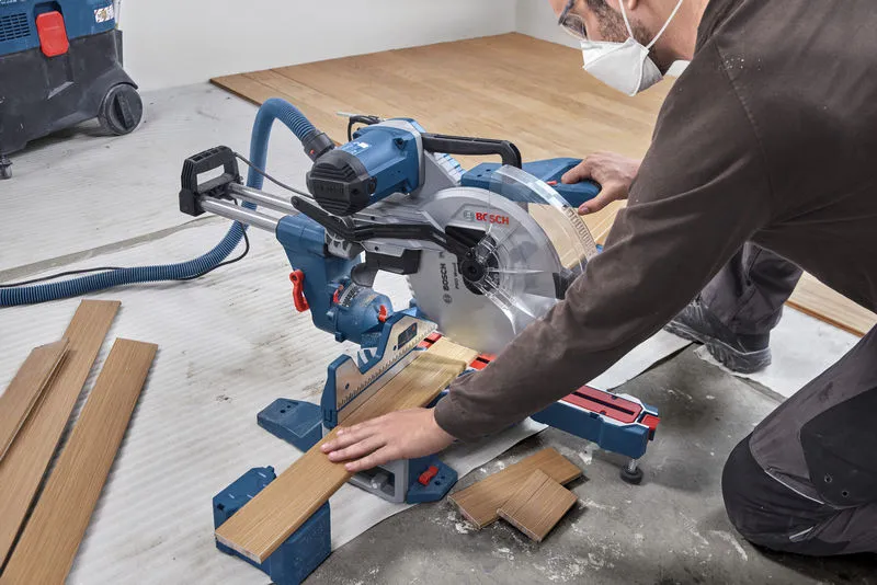 A person wearing safety equipment cuts wooden flooring with a sliding mitre saw.