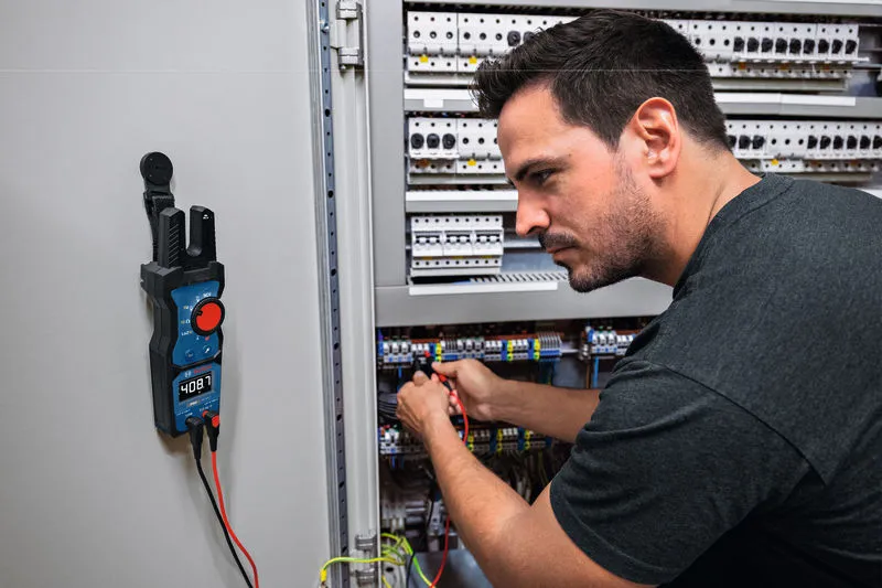 A person tests electrical connections in a control cabinet using a digital multimeter.