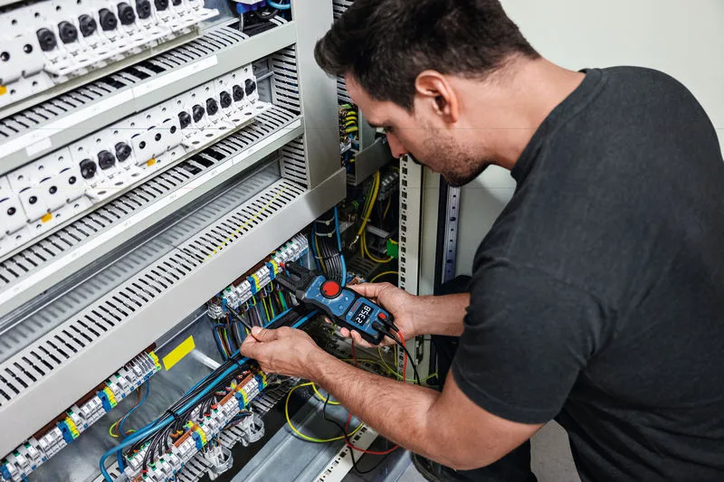A person measures electrical values in a control panel using a fork meter.