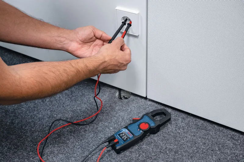 Person testing a wall socket voltage with a digital clamp meter on a carpeted floor.