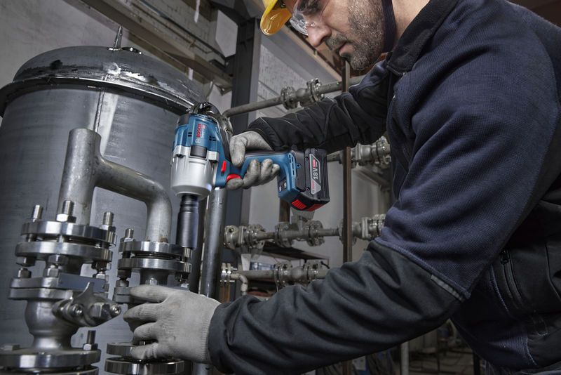 A person wearing safety equipment uses an impact wrench to fasten a pipe flange.