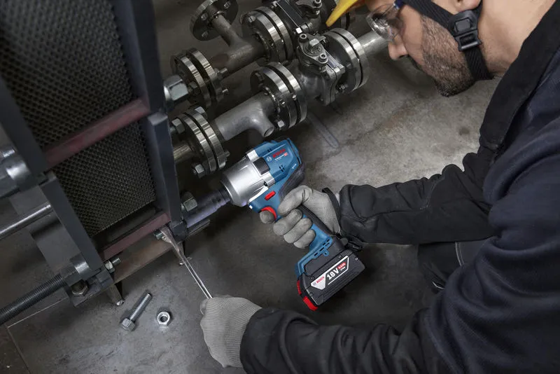 A person wearing safety equipment uses a cordless impact wrench on industrial machinery.
