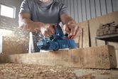 A person wearing safety equipment planes a wooden beam, wood shavings flying.