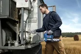 A person wearing safety equipment uses a power tool to service farm machinery in a field.