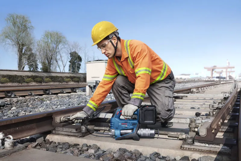 A worker wearing safety equipment uses a power tool to tighten bolts on railway tracks.